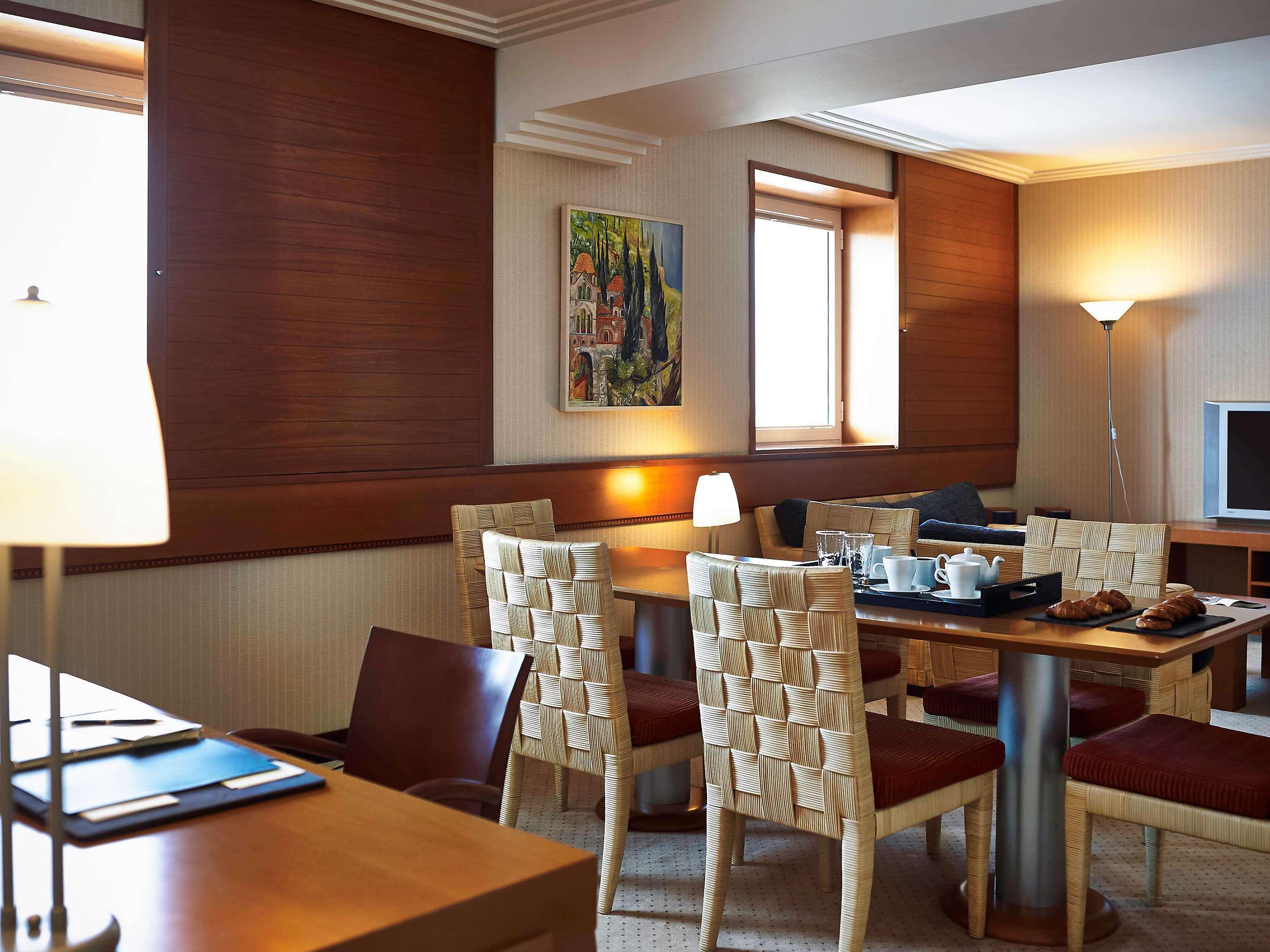 فندق سوفيتِل أثينا إيربورت Spáta المظهر الخارجي الصورة The photo shows a cozy dining area, likely in a hotel or a modern apartment. There is a wooden table surrounded by four chairs with woven backs. On the table, there's a tray with breakfast items, including some pastries. The walls are adorned with wa