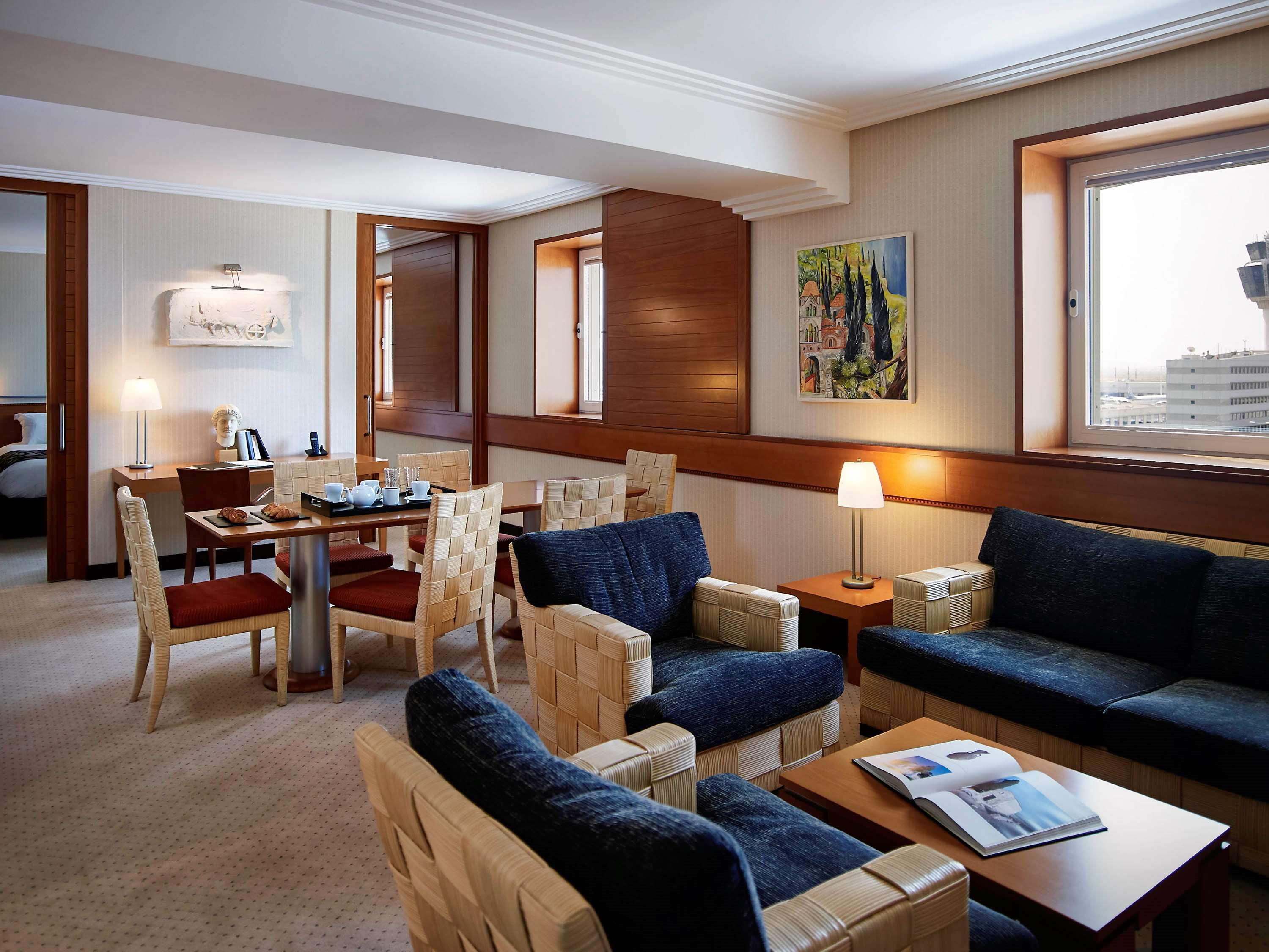 فندق سوفيتِل أثينا إيربورت Spáta المظهر الخارجي الصورة The photo shows a modern living area of a hotel suite. The room features a comfortable seating arrangement with dark blue sofas and armchairs, along with a wooden coffee table. There is a dining area with a table and chairs adjacent to the seating ar