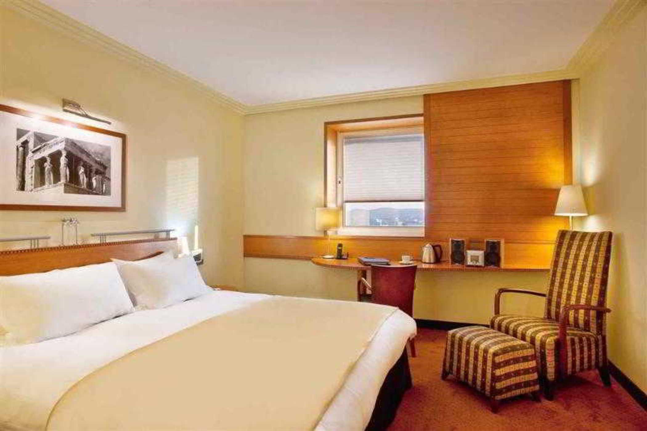 فندق سوفيتِل أثينا إيربورت Spáta المظهر الخارجي الصورة The photo depicts the interior of a hotel room. It features a large bed with white linens and a beige blanket. There is a window with blinds, allowing some natural light to enter the space. Beside the bed, there is a bedside lamp and a piece of artwo