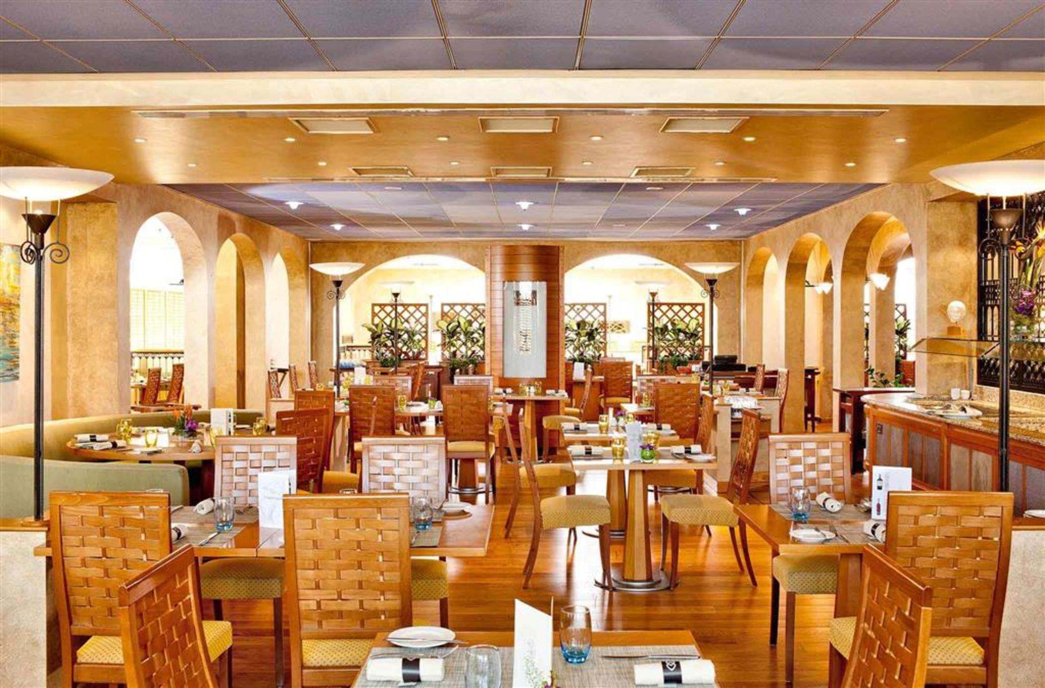 فندق سوفيتِل أثينا إيربورت Spáta المظهر الخارجي الصورة The photo shows the interior of a restaurant. The space features warm, golden tones with wooden furniture. There are several tables with chairs arranged throughout the room, and the tables appear to be set for dining with some plates and utensils. Ar