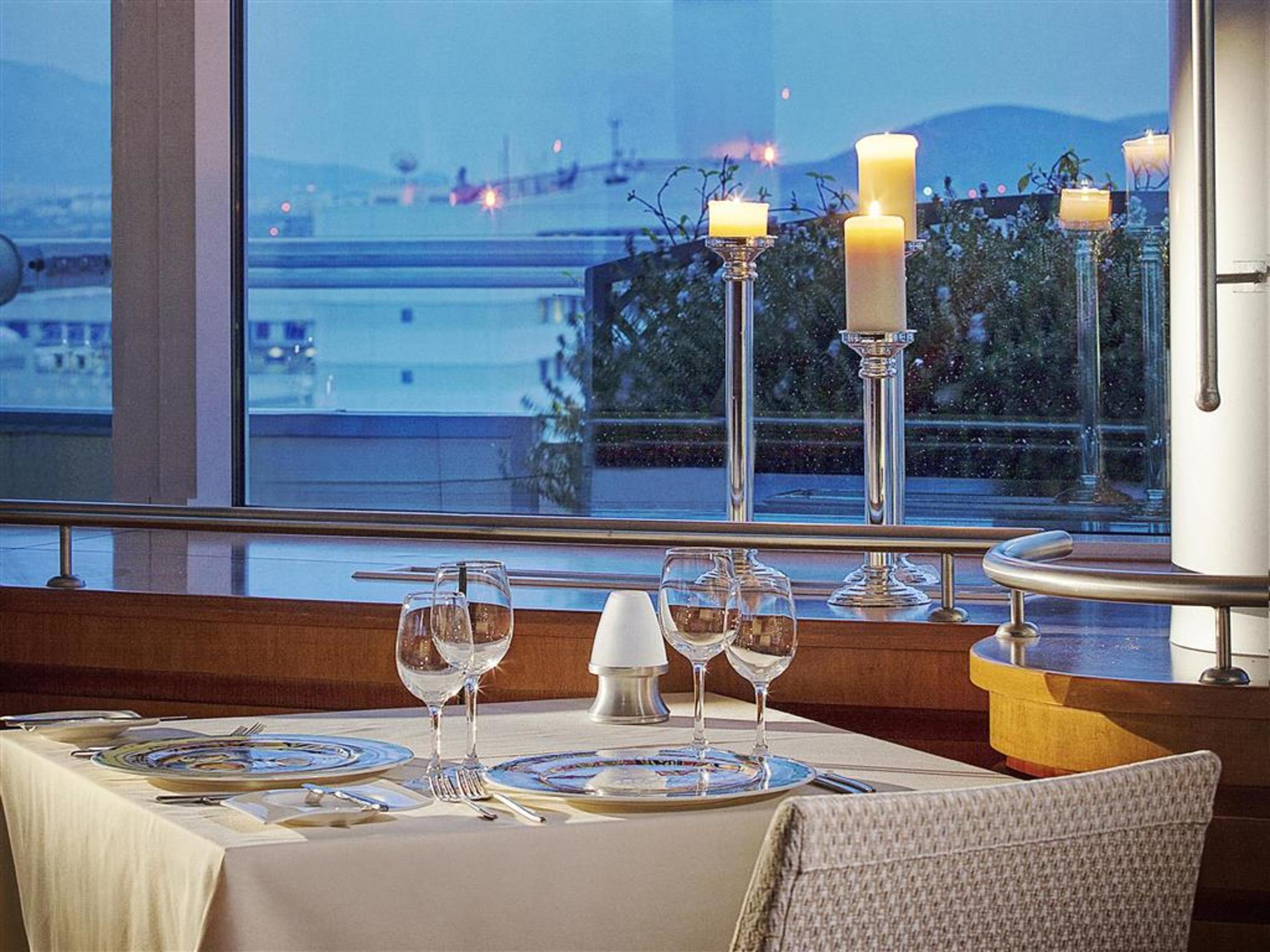 فندق سوفيتِل أثينا إيربورت Spáta المظهر الخارجي الصورة The photo shows an elegant dining setup in a restaurant. The table is covered with a light-colored tablecloth and is beautifully arranged with glassware, including wine glasses and a small salt shaker. In the background, there are large windows that 