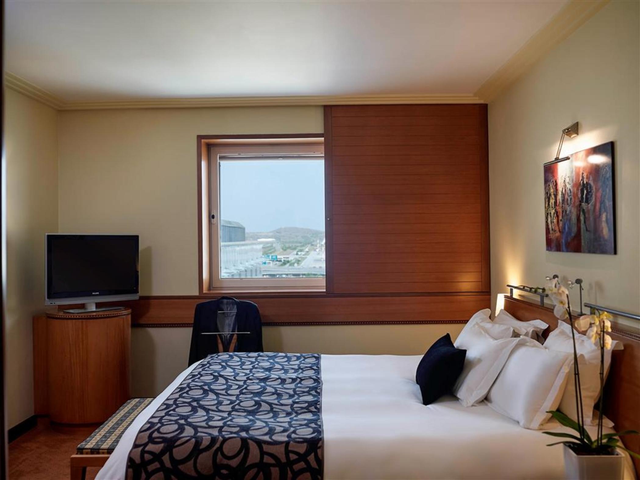 فندق سوفيتِل أثينا إيربورت Spáta المظهر الخارجي الصورة The photo depicts a cozy hotel room featuring a large bed with white linens and a patterned blue throw. There are several pillows on the bed, and a bedside table with a small lamp. A window allows natural light to enter the room, and it has wooden bl