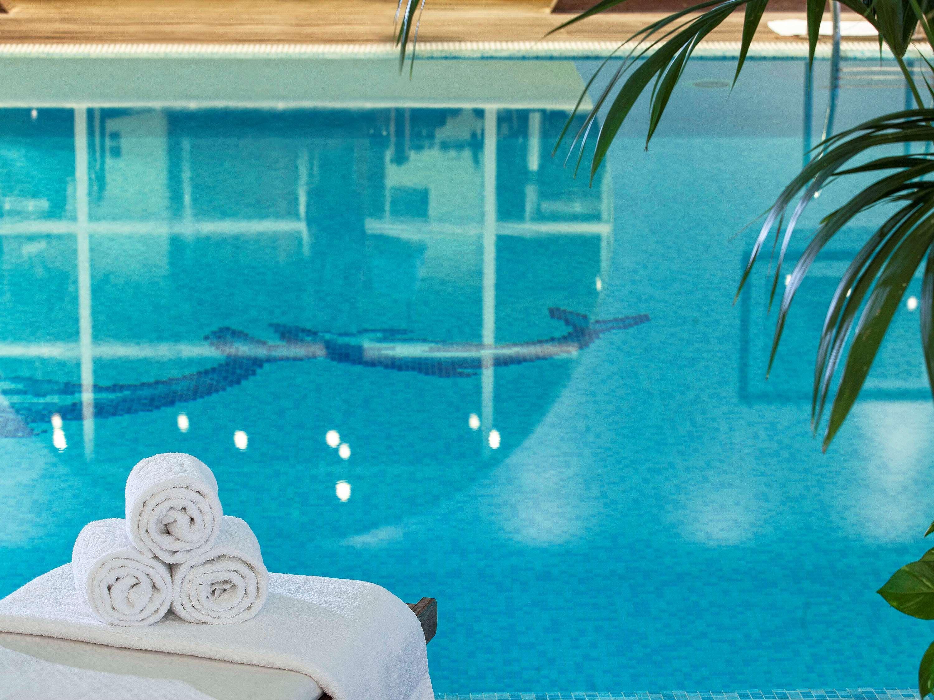 فندق سوفيتِل أثينا إيربورت Spáta المظهر الخارجي الصورة The photo shows a serene swimming pool with clear, turquoise water. In the foreground, there are neatly rolled towels placed on a lounge chair. The water reflects soft lighting and has a tiled pattern visible beneath the surface, while there are hint