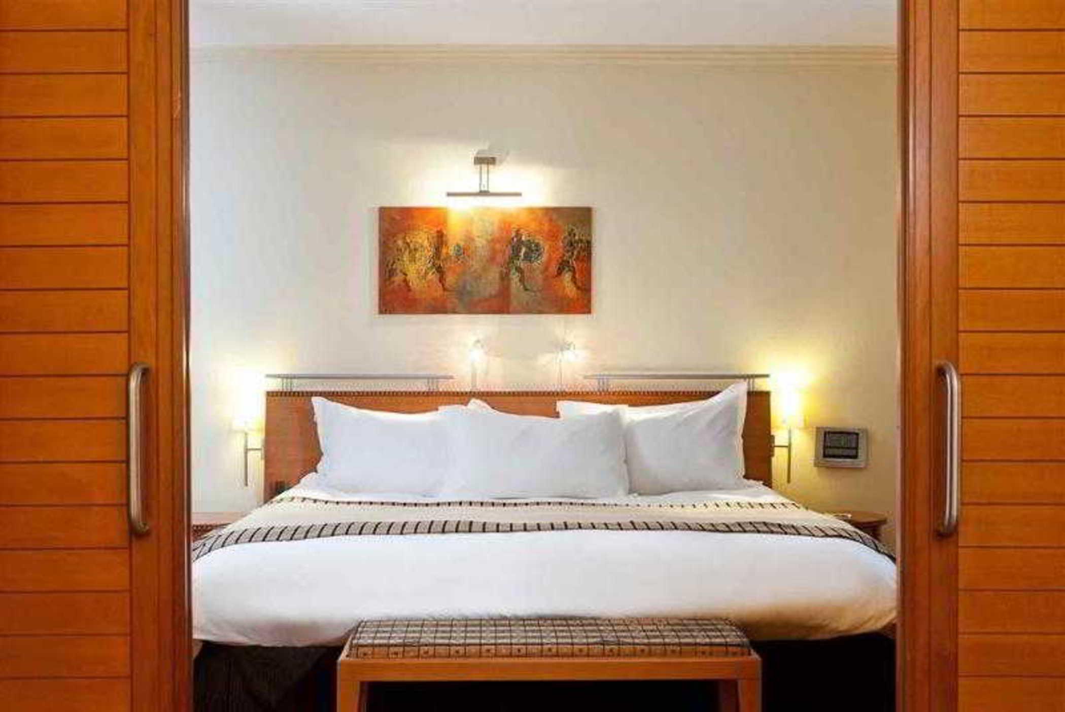 فندق سوفيتِل أثينا إيربورت Spáta المظهر الخارجي الصورة The photo shows a well-decorated hotel room with a contemporary design. At the center, there is a large bed with a white comforter and several pillows. Behind the bed, there is a piece of artwork featuring figures, which adds a pop of color to the ro