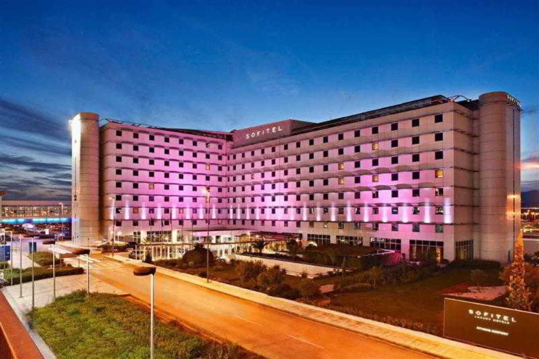 فندق سوفيتِل أثينا إيربورت Spáta المظهر الخارجي الصورة The photo displays a modern hotel building, likely a hotel brand, with a sleek, contemporary architectural design. The façade is illuminated in pink and purple lights, suggesting a vibrant atmosphere. The building has multiple floors, with large wind