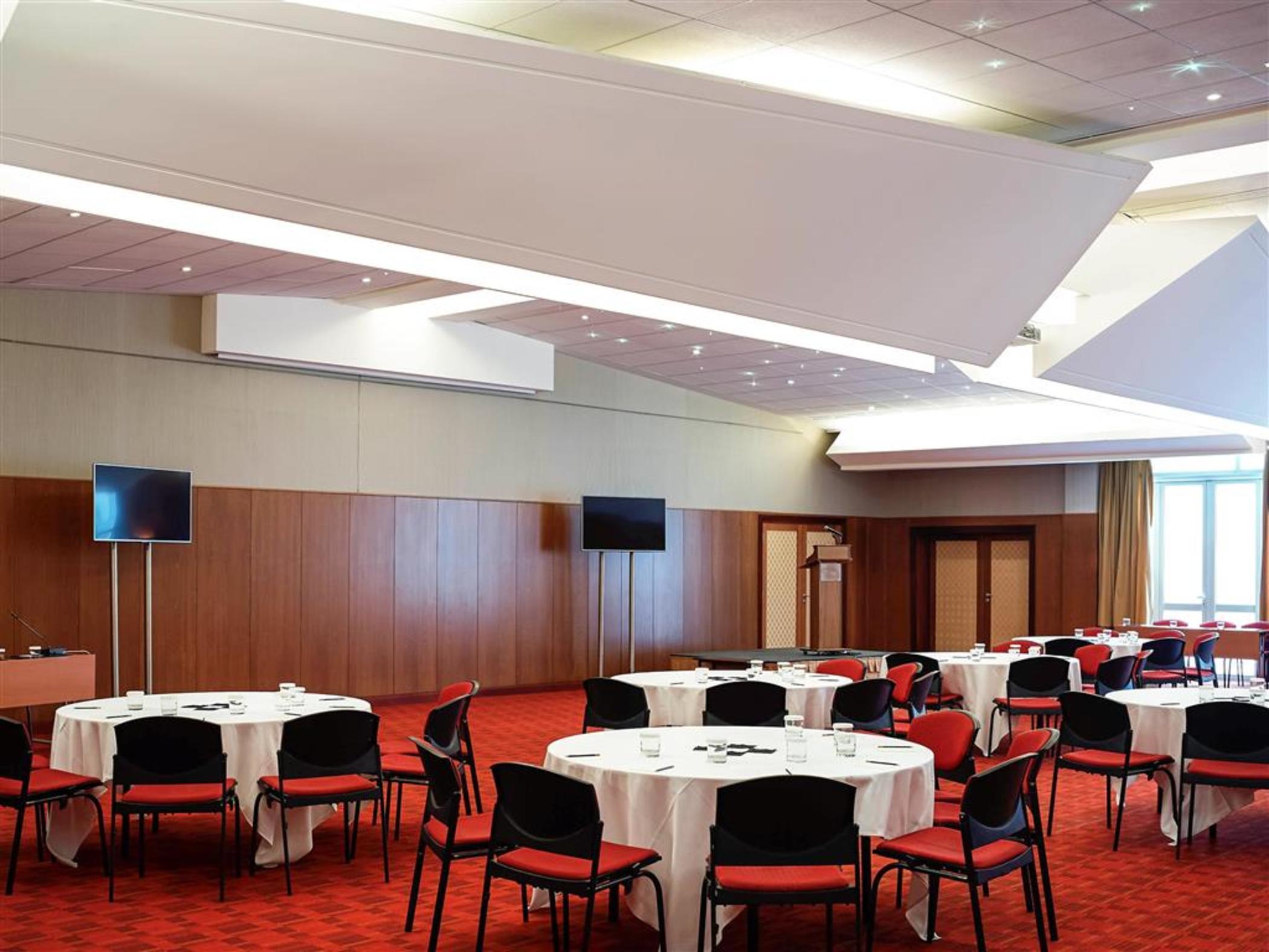فندق سوفيتِل أثينا إيربورت Spáta الاعمال الصورة The photo depicts an indoor event space, likely set up for a conference or gathering. The room features several round tables with white tablecloths and black chairs, arranged neatly across the floor. The flooring is carpeted in a red pattern, adding 
