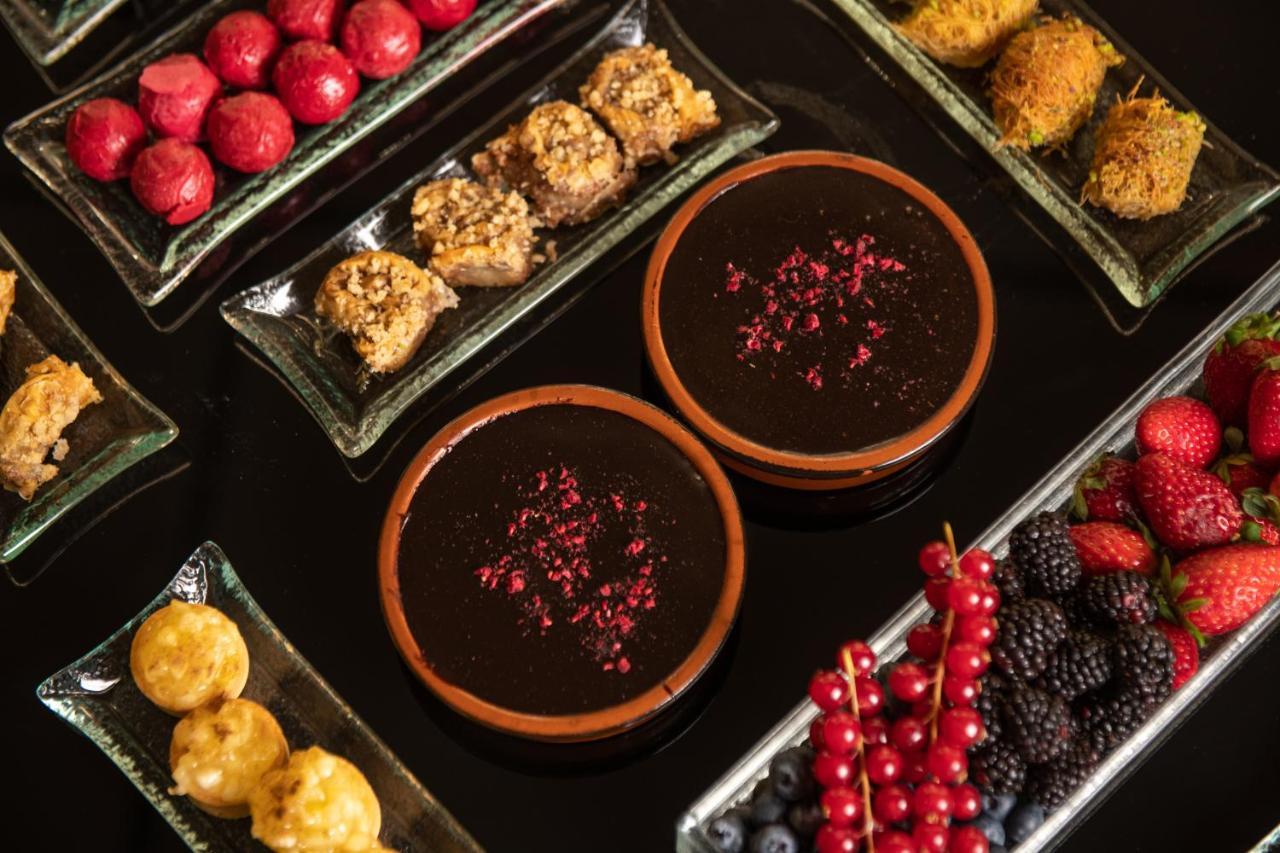 فندق سوفيتِل أثينا إيربورت Spáta المظهر الخارجي الصورة The image shows a beautifully arranged assortment of desserts and sweets on a dark surface. There are two brown bowls filled with a thick, dark dessert, garnished with rose petals. Surrounding the bowls are various plates containing a variety of trea