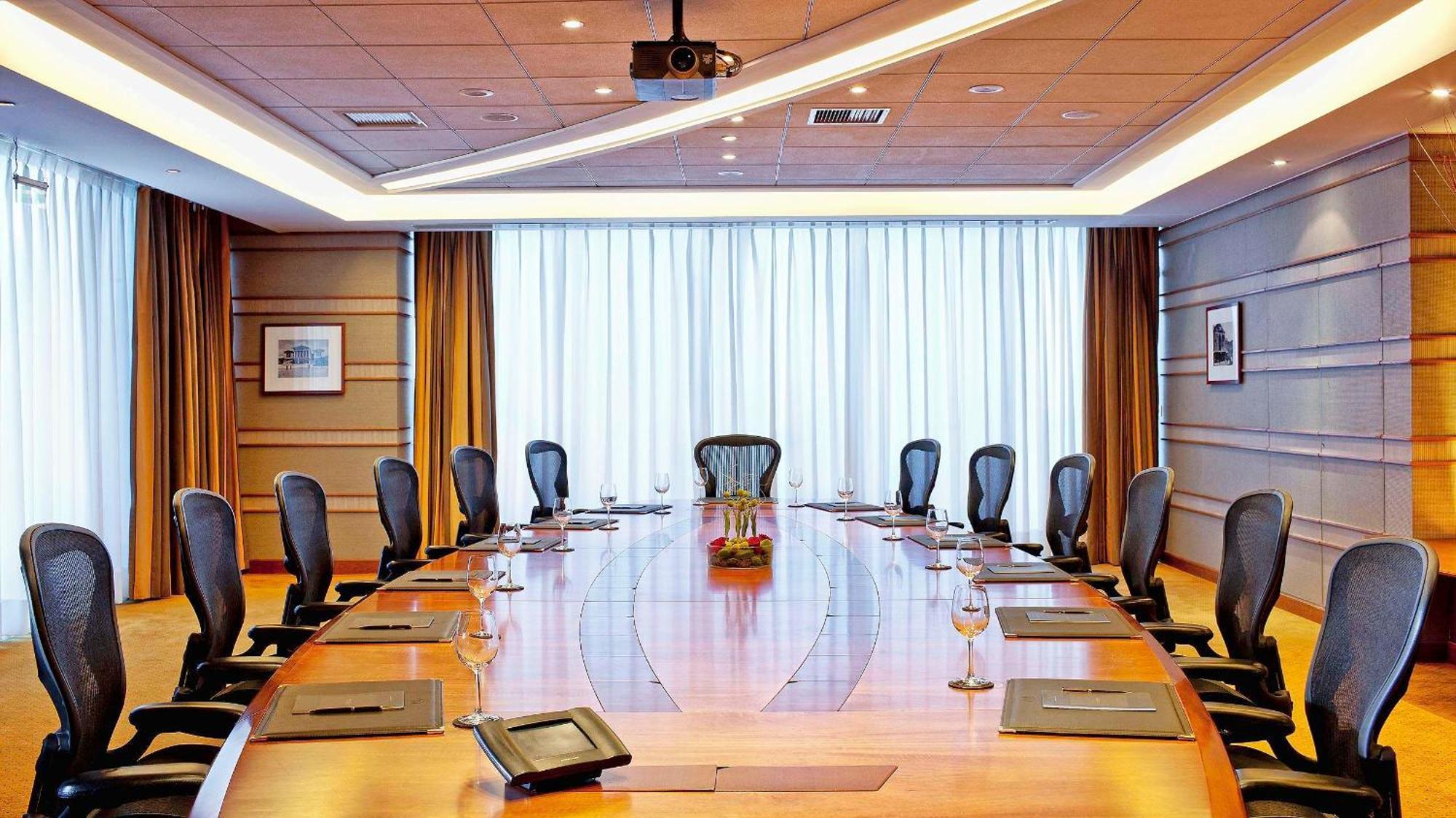 فندق سوفيتِل أثينا إيربورت Spáta المظهر الخارجي الصورة The photo depicts a modern conference room featuring a large, oval wooden table surrounded by several ergonomic chairs. The room has dim lighting and is illuminated by natural light coming in through large windows covered by sheer curtains. There are