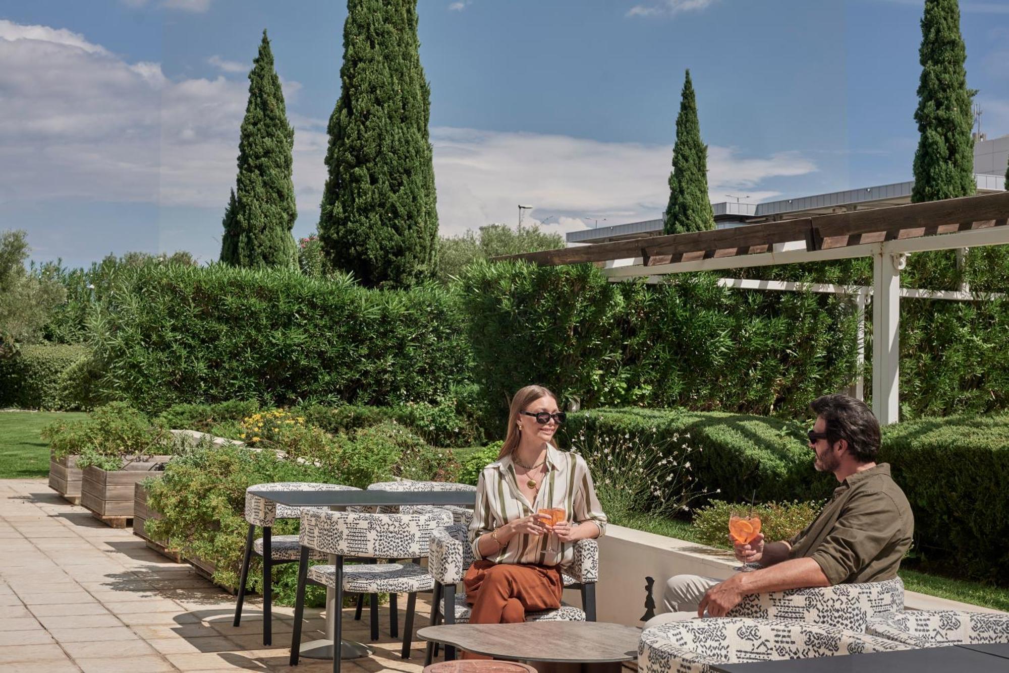 فندق سوفيتِل أثينا إيربورت Spáta المظهر الخارجي الصورة The photo shows two people seated at an outdoor setting, enjoying drinks. They are positioned at a table with decorative tablecloths, surrounded by lush greenery and tall cypress trees. The atmosphere appears relaxed and sunny, suggesting a pleasant 