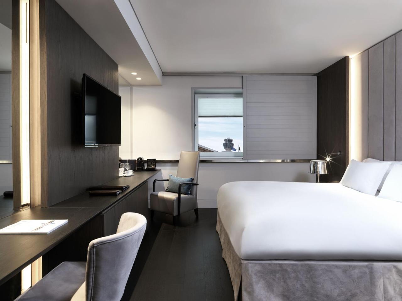 فندق سوفيتِل أثينا إيربورت Spáta المظهر الخارجي الصورة The photo shows a modern hotel room. It features a large bed with white bedding, a sleek desk with a chair, and a flat-screen TV mounted on the wall. There are large windows allowing natural light to enter, and the room is decorated in neutral tones 